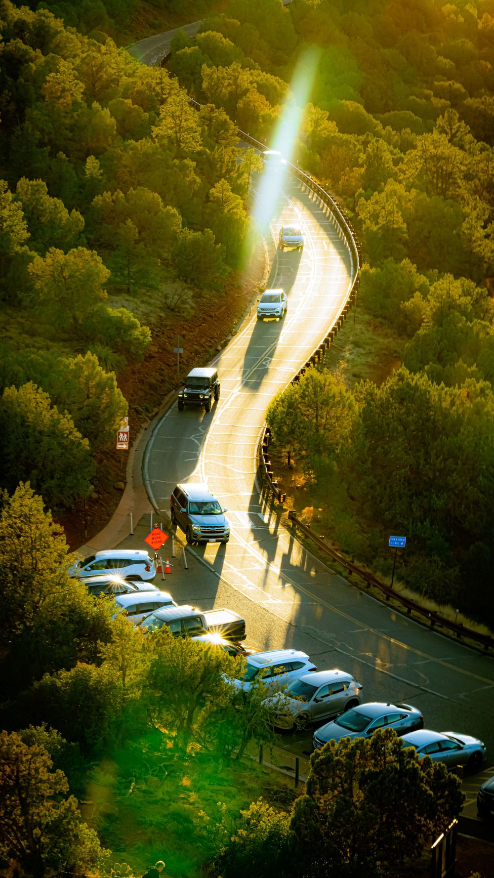 Curved mountain road with parked cars and dramatic sunlight streaming through dense forest.