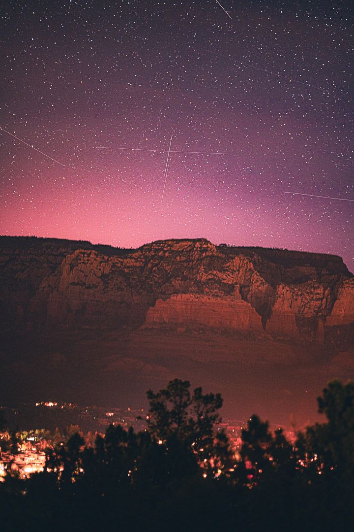 Starry night sky over red rock cliffs with city lights glowing at the base of canyon walls.