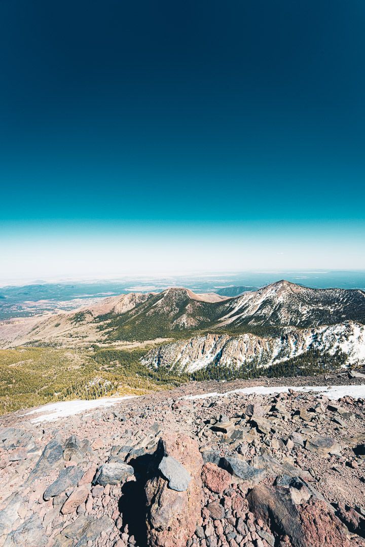 Rocky mountain summit with scattered boulders and distant peaks under a bright azure sky.