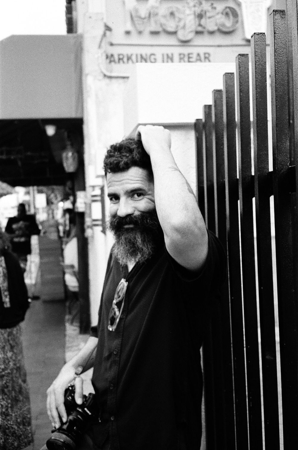 Black and white street photo showing a bearded man in dark clothing leaning against metal bars outside a storefront.