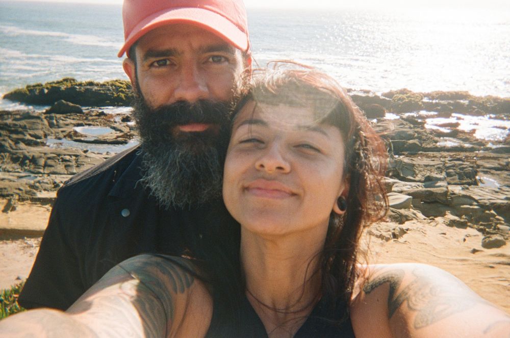 A loving couple takes a sunny selfie together on a rocky oceanside beach during a bright afternoon.