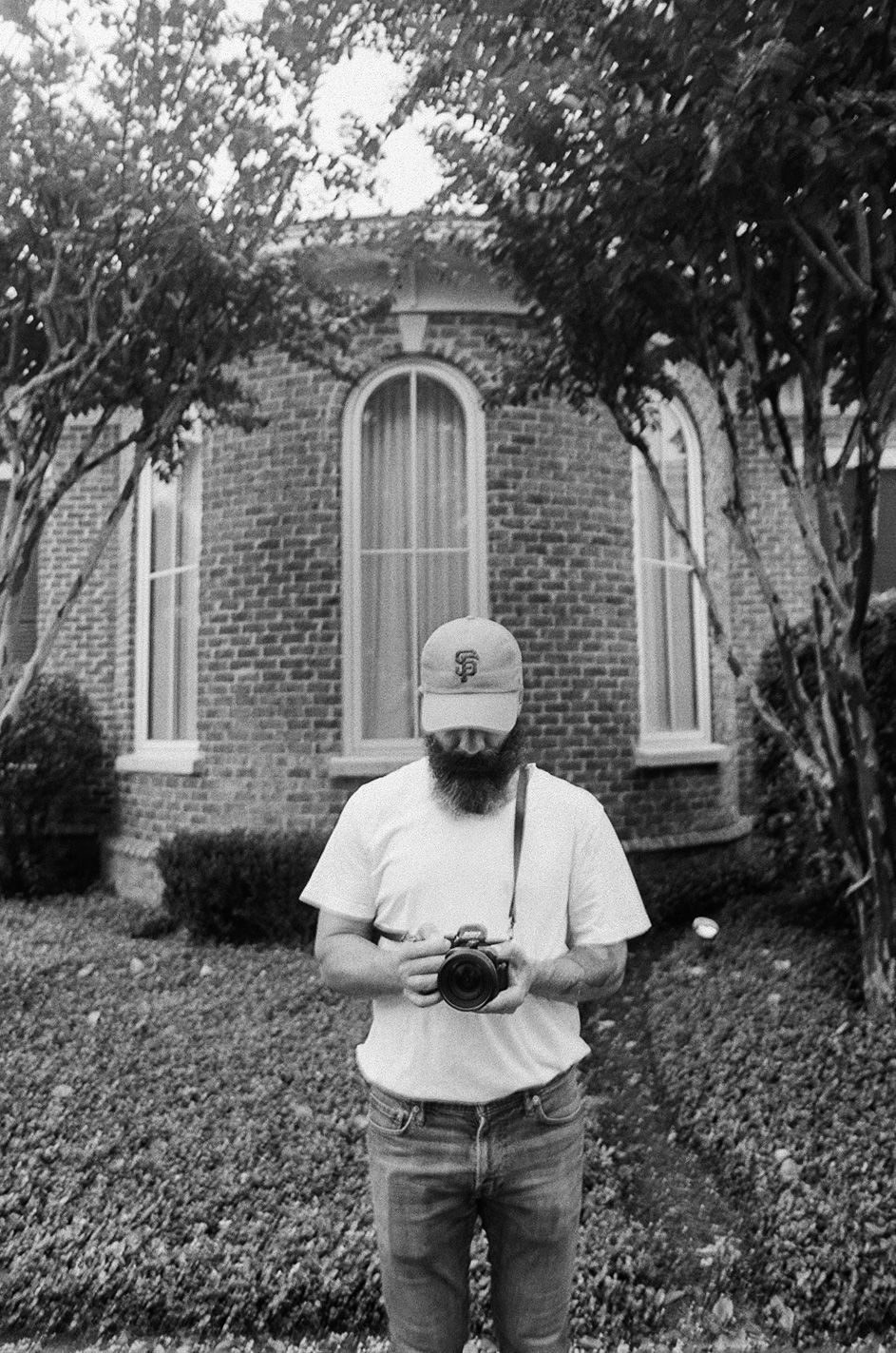 Black and white photo of someone holding a camera outside a brick church with arched windows and surrounding landscaping.