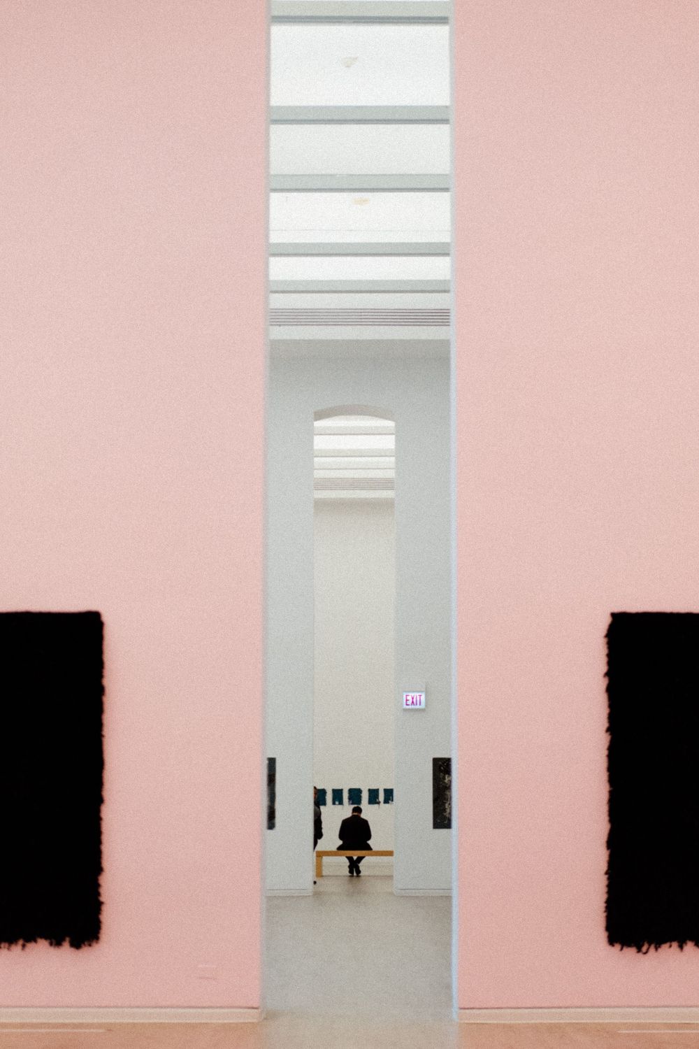 Minimalist art gallery with pink walls, black paintings and a distant solitary figure seated in a white archway.