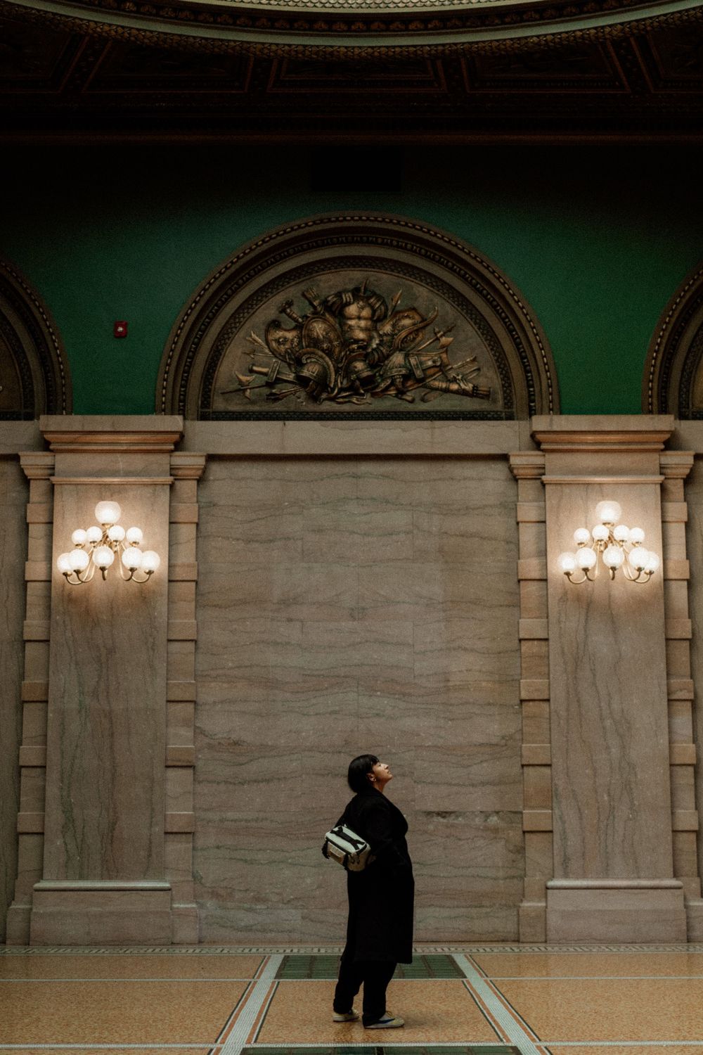 A person in black clothing stands before an ornate marble wall with grand columns and illuminated sconces in a classical interior.