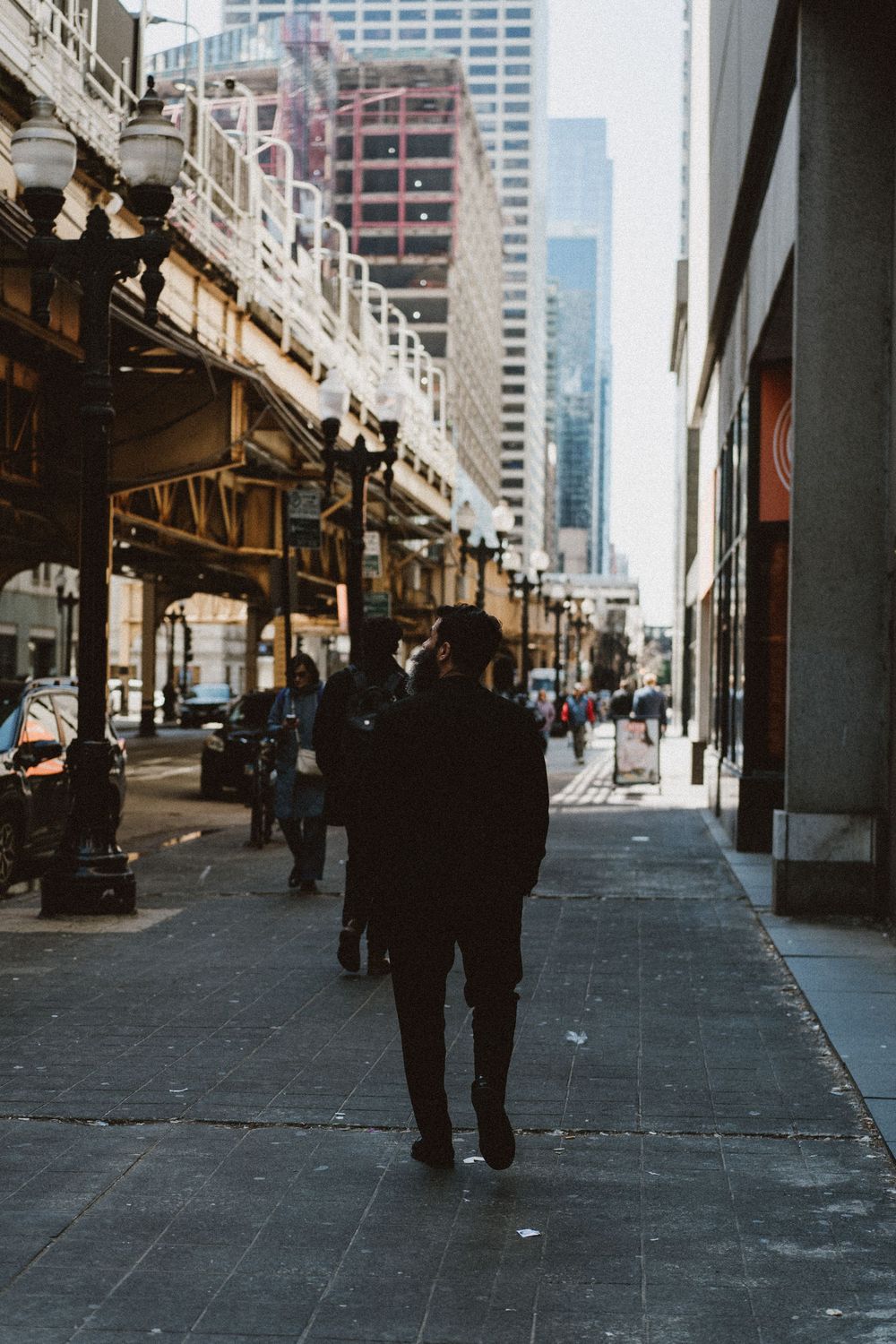 Urban street scene in Chicago with elevated train tracks above, pedestrians walking beneath skyscrapers on a city sidewalk.