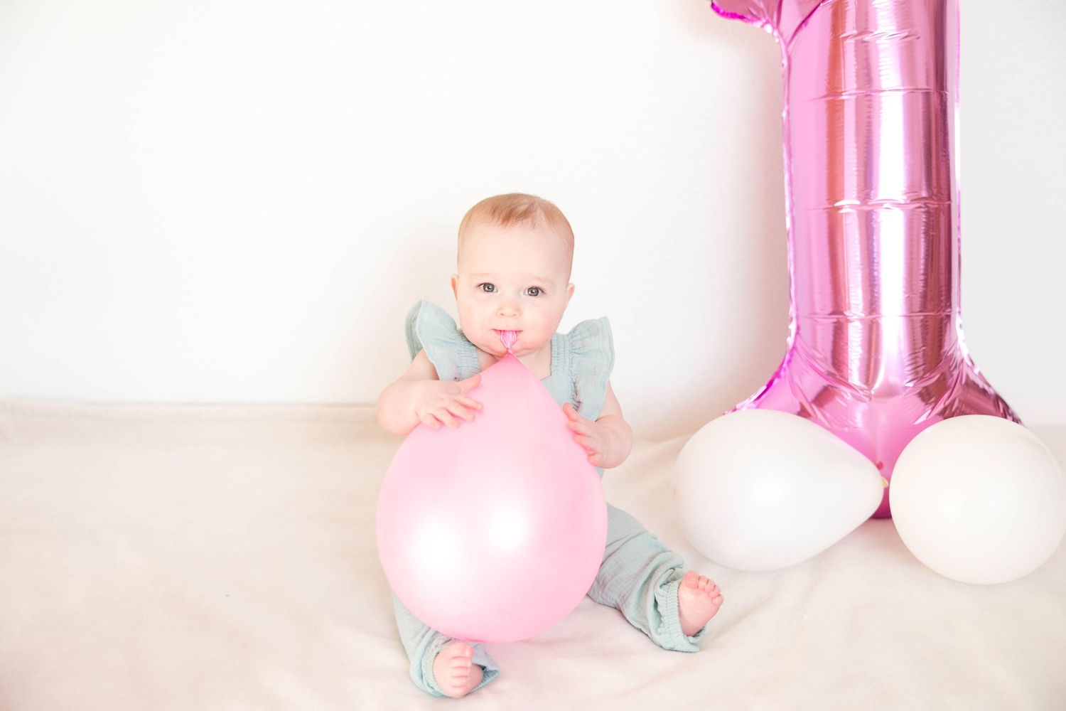Baby in gray outfit plays with pink balloon next to party decorations.