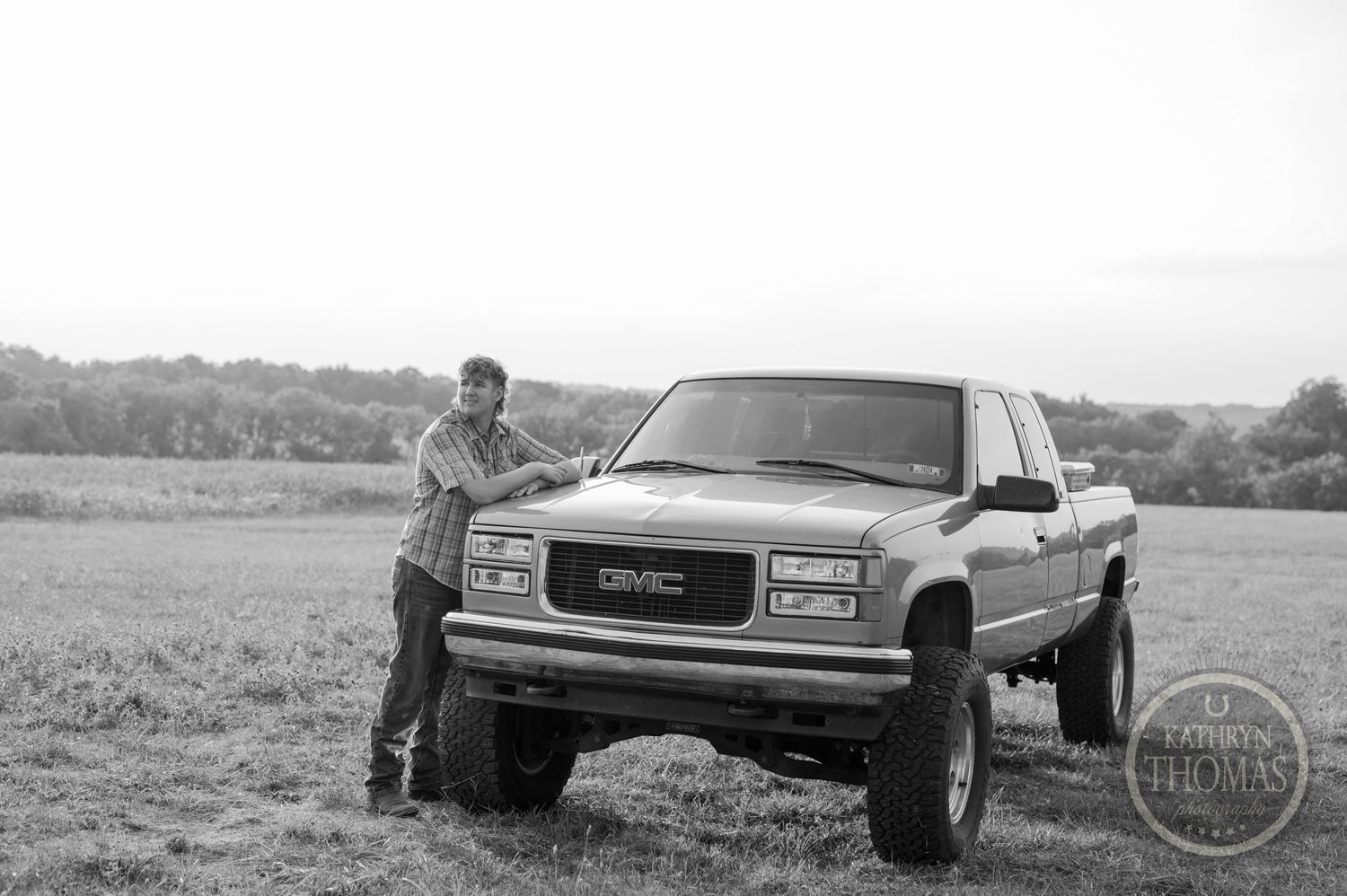 Black and white photo of lifted GMC Sierra truck with someone leaning against it in field.