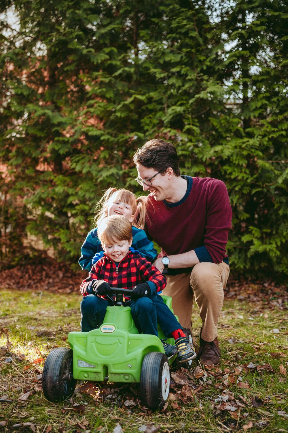 Playful moment outdoors as parent helps child ride a green toy pedal tractor on fallen autumn leaves.