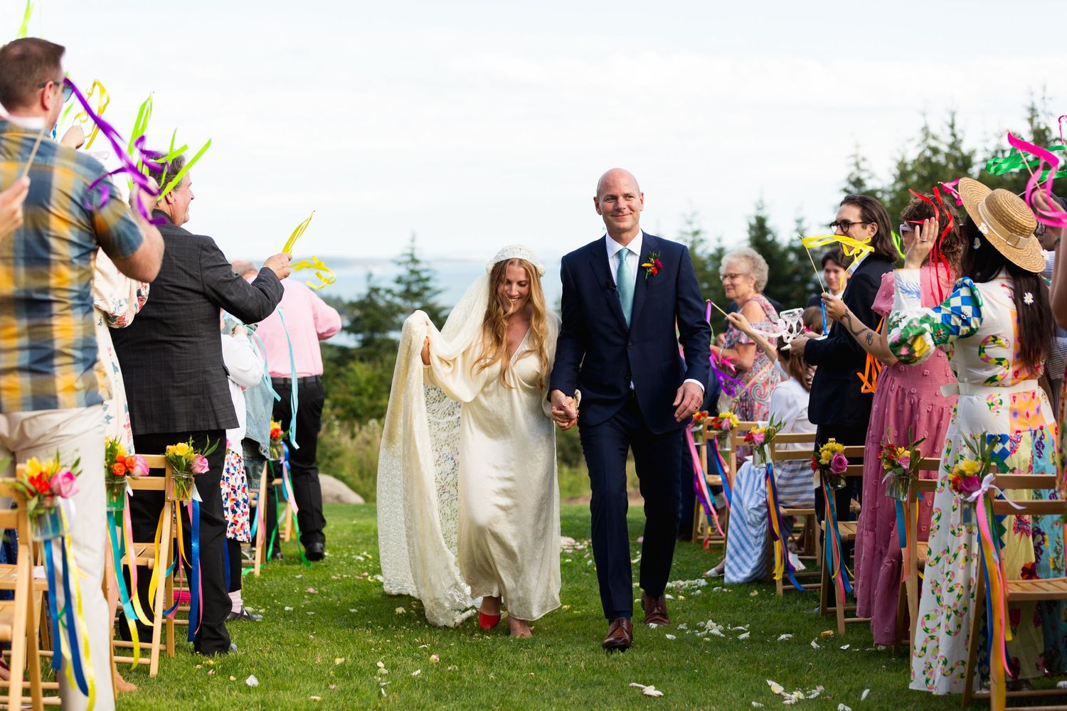 Wedding couple walks down aisle while guests throw colorful streamers and confetti during outdoor ceremony.