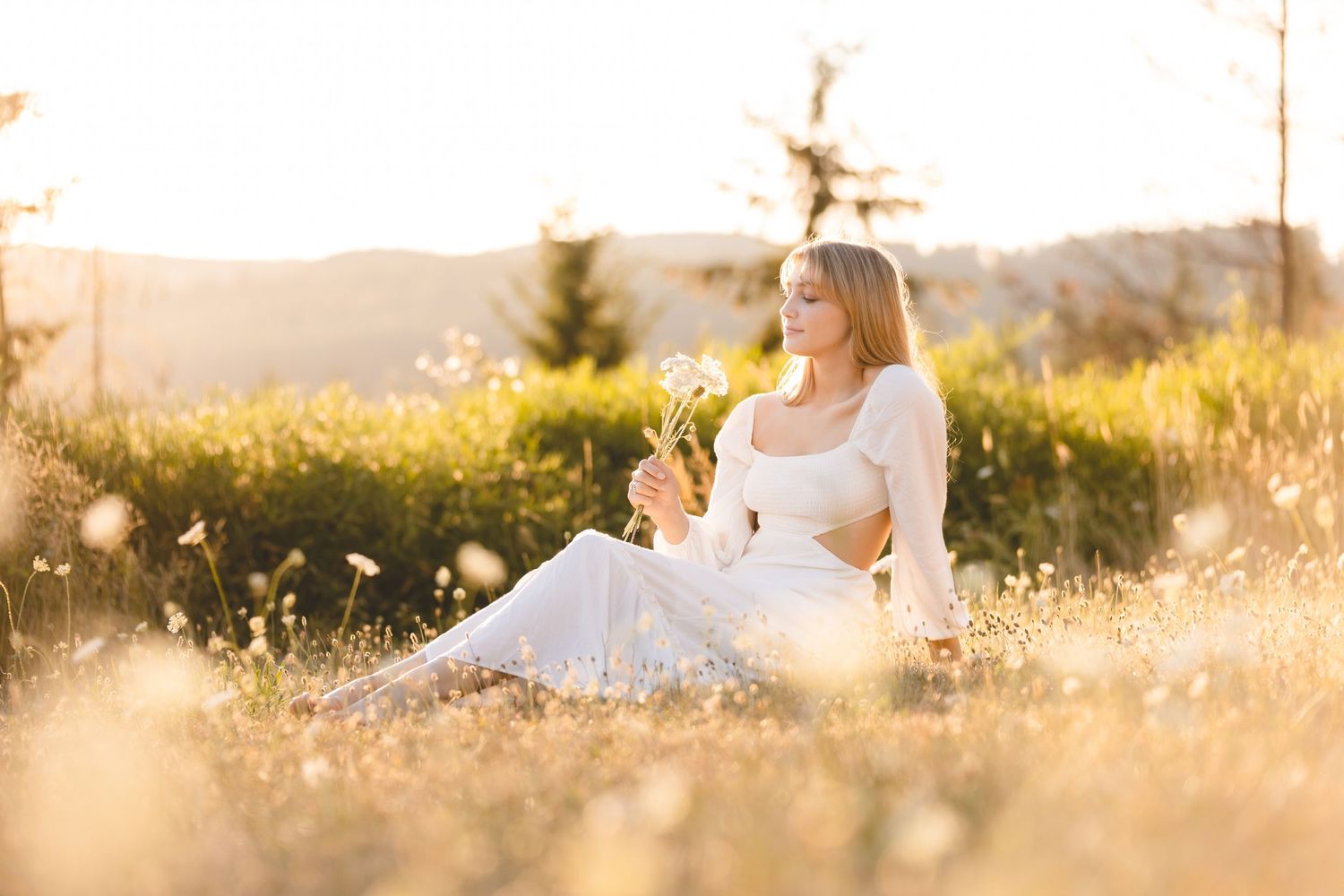 Person in white dress sits peacefully in golden sunlit meadow with wildflowers at sunset.