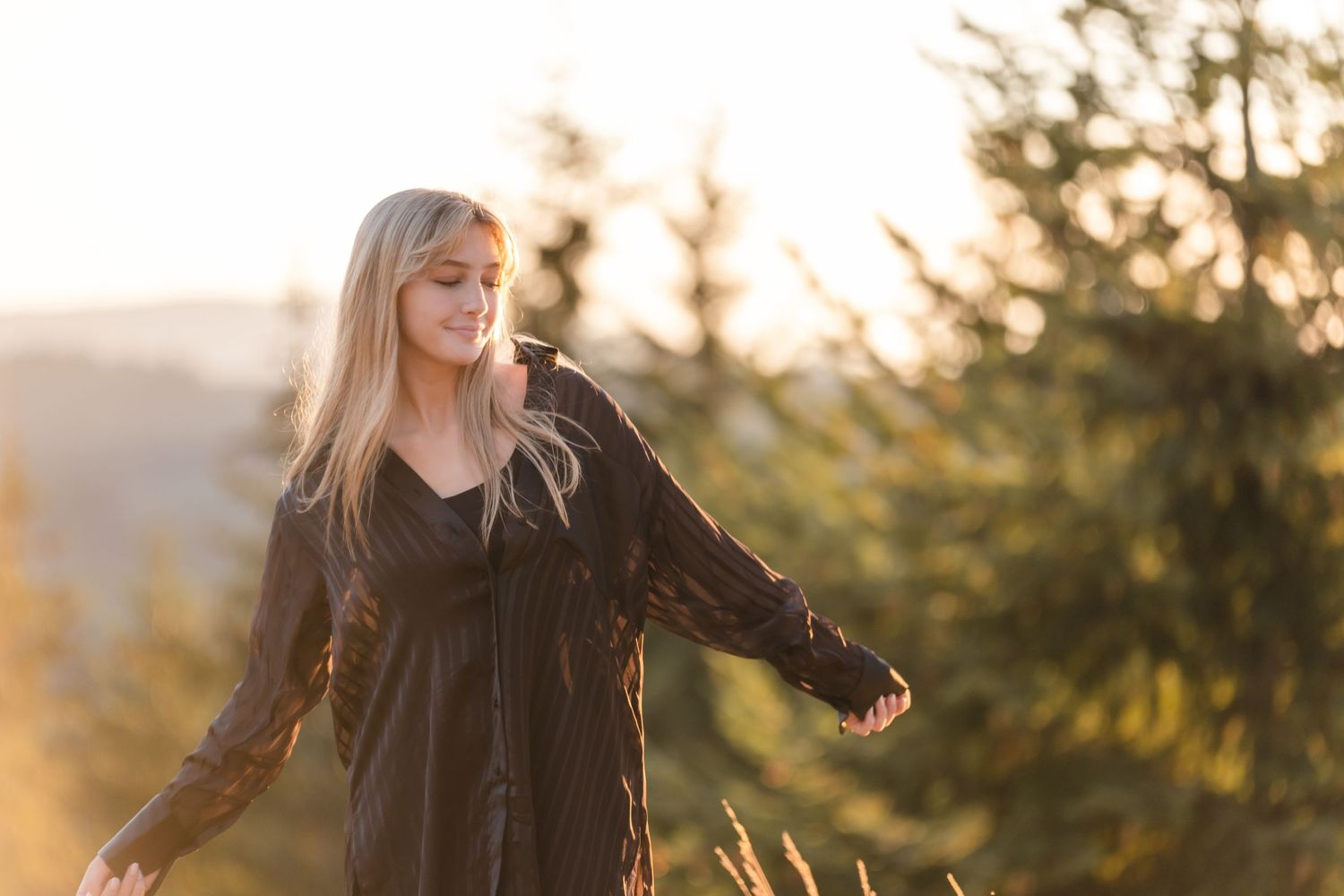 Woman in black dress poses in golden sunlight against blurred forest background at sunset.