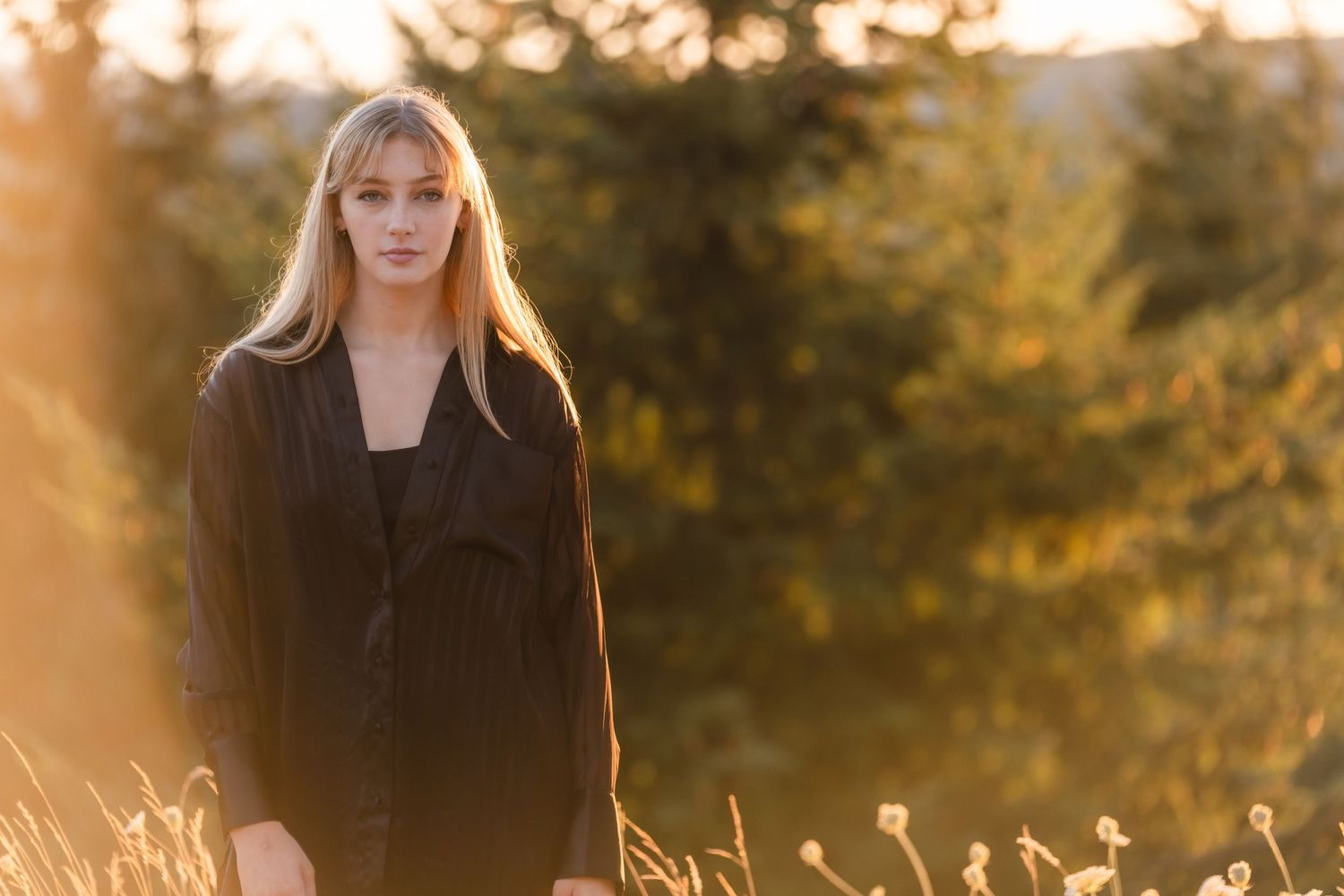 Person in a black coat standing in a sunlit field with golden light filtering through fall foliage at sunset.