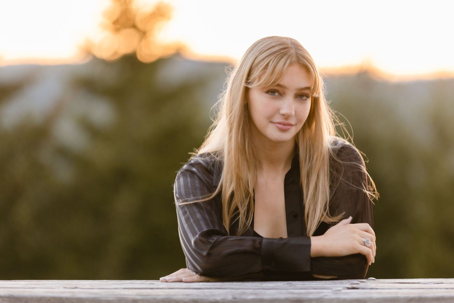 A woman in a black leather jacket leans on a wooden railing during golden hour sunset with mountains in background.
