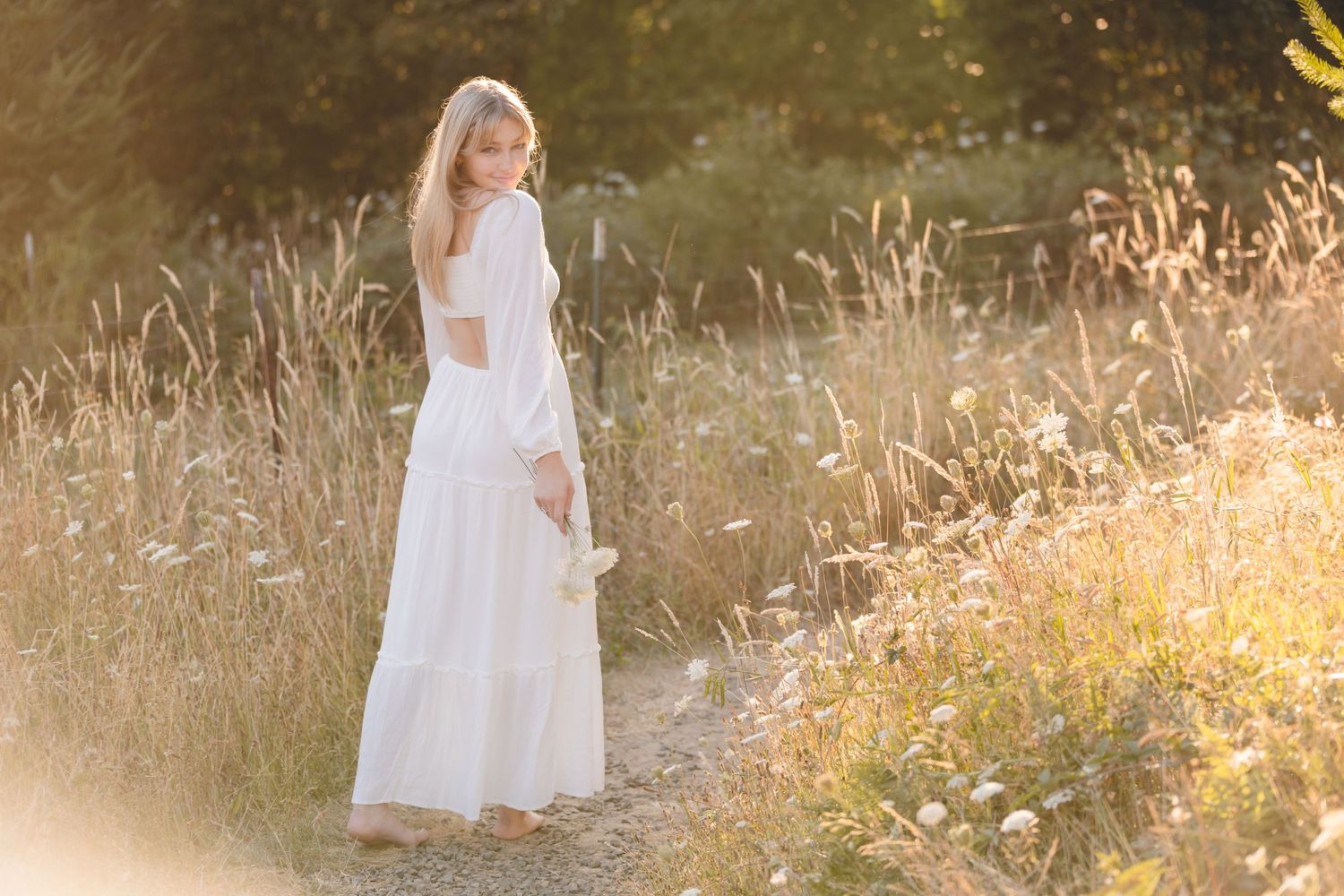 A person wearing a long white dress stands in a golden sunlit meadow during a summer evening.