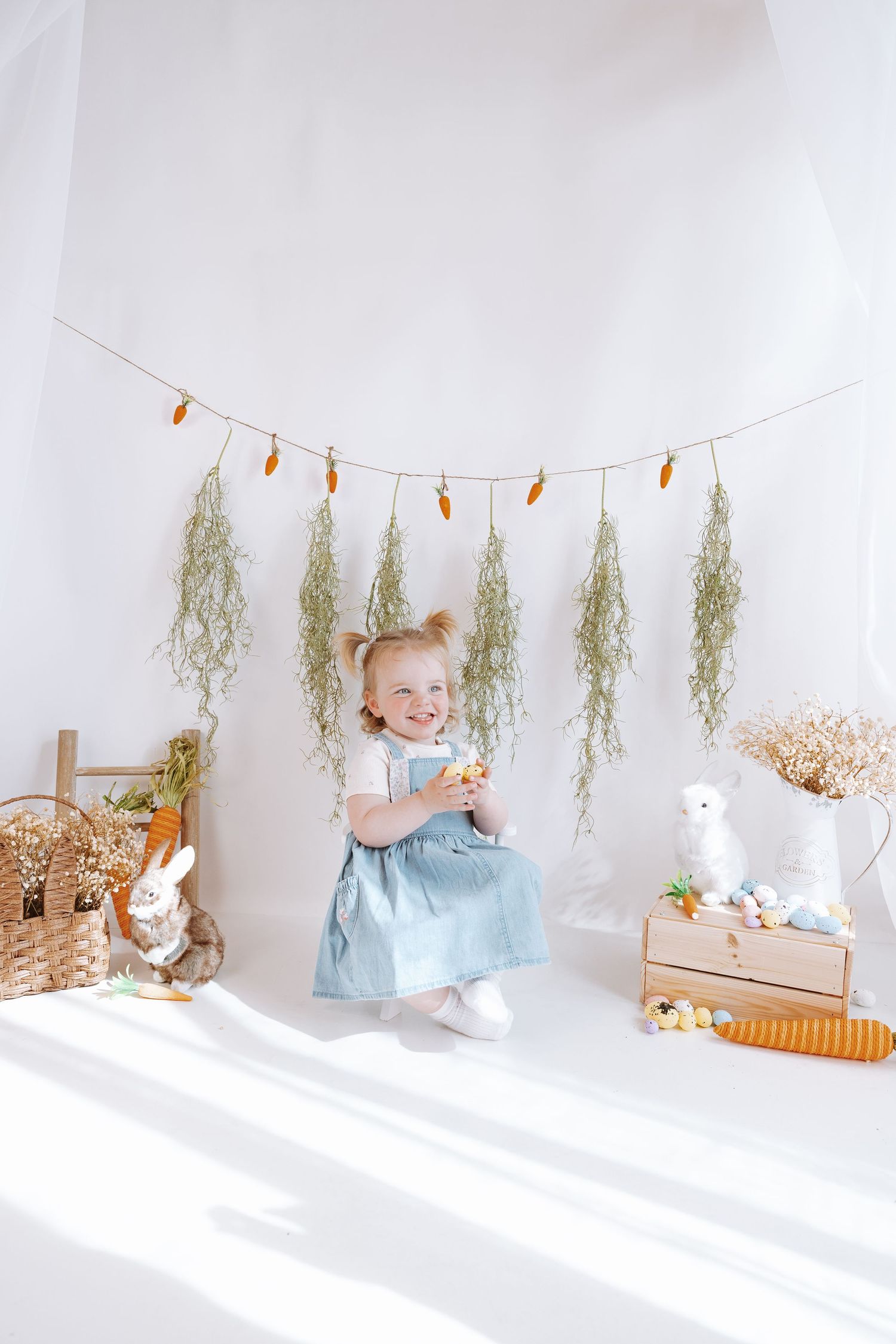 Light and airy Easter photo setup with hanging dried flowers, bunny decorations, and a small child in blue dress.