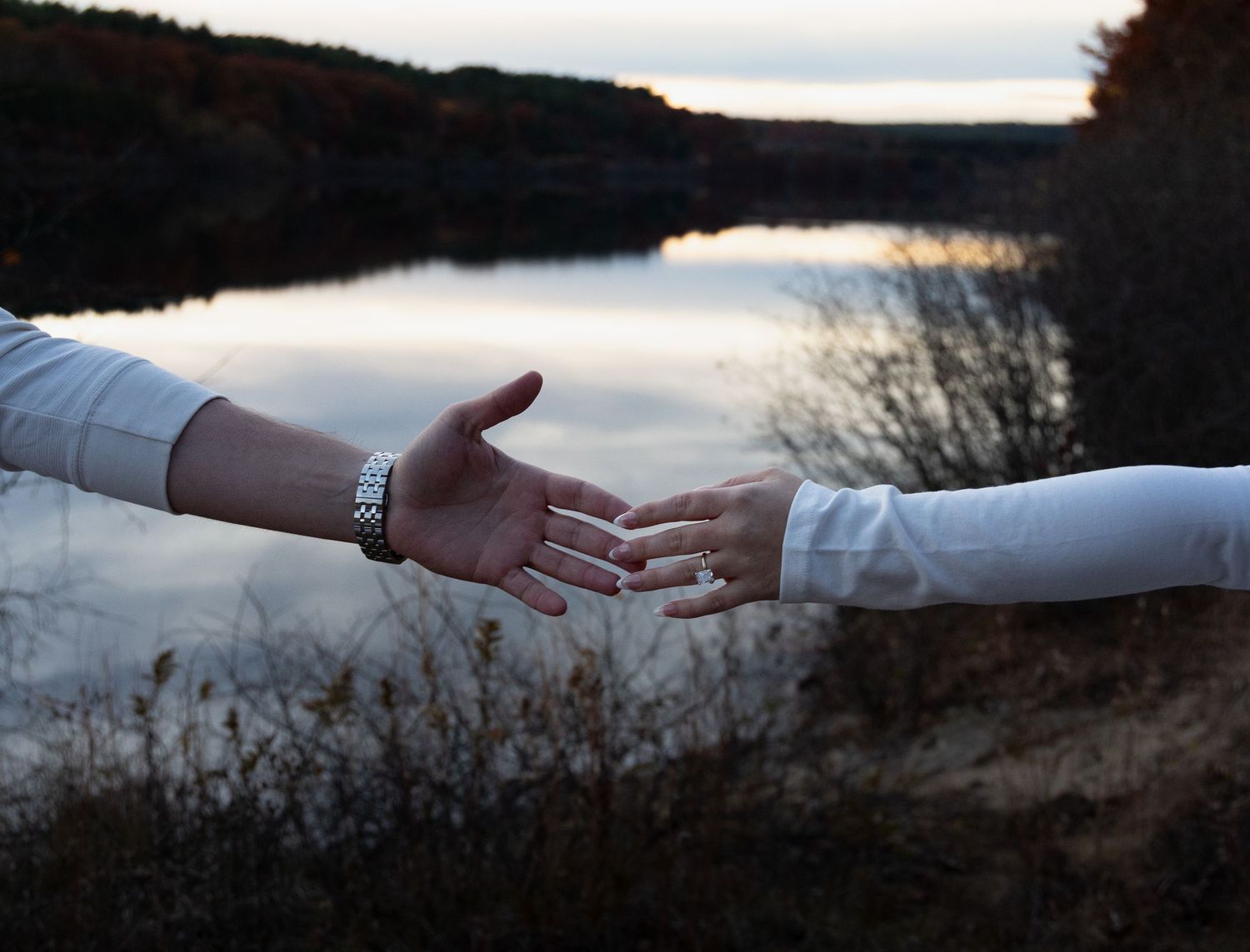 Two people reaching toward each other over scenic lake at dusk with autumn landscape background.