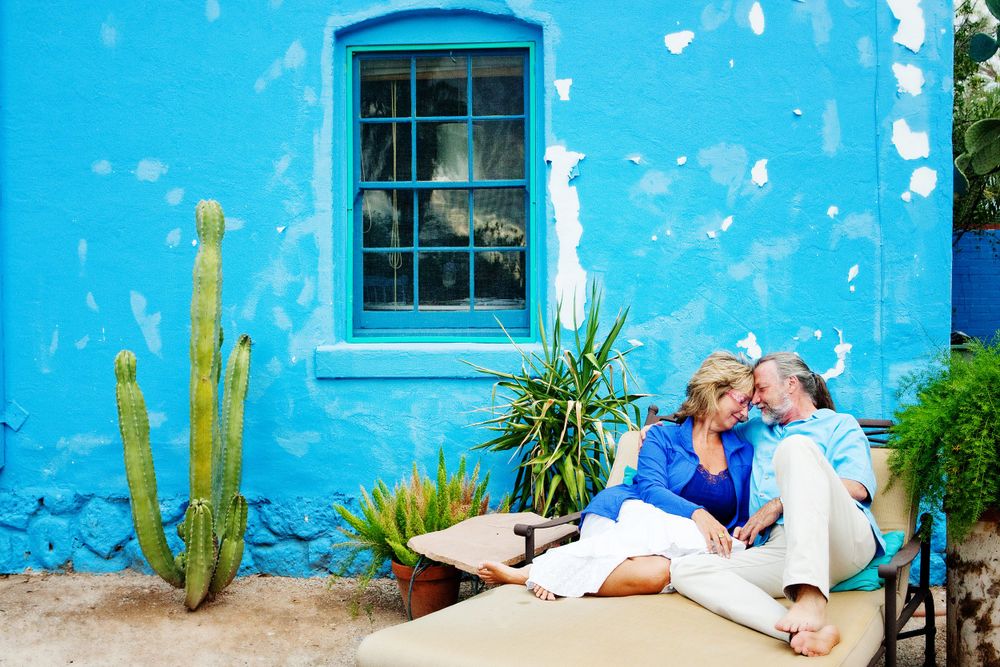 A blue-washed adobe home in Tucson with a window frames desert plants and people lovingly embracing.