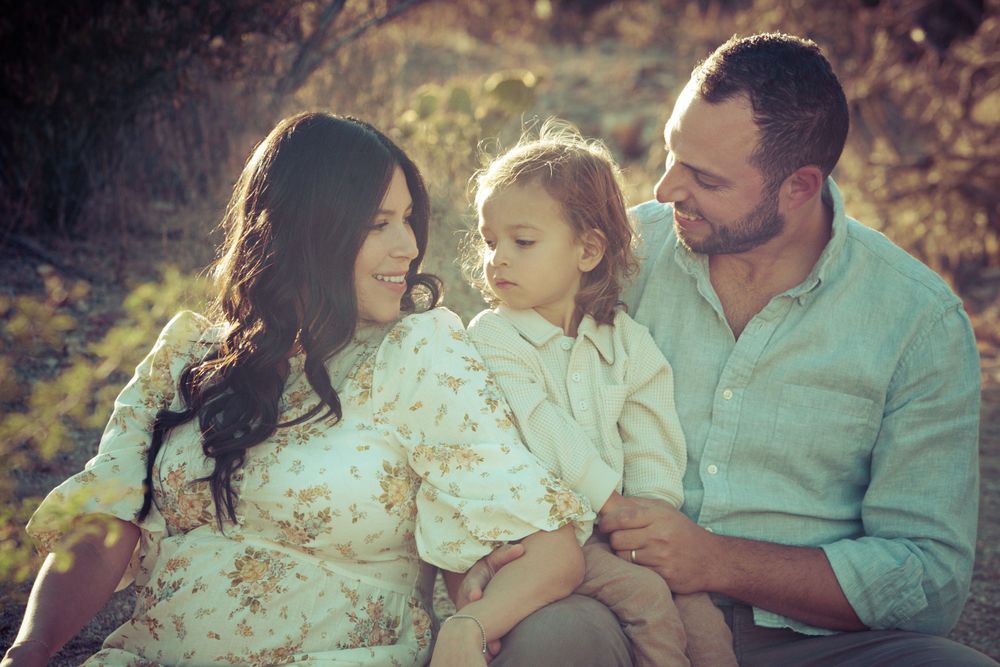 A  young family with a two year old boy sits together outdoors as golden sunlight filters through the background in Saguaro National Park.