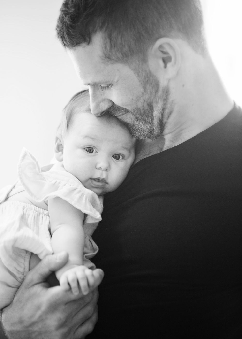 A tender black and white profile portrait shows a Dad cradling a two week old baby girl wearing a ruffled outfit.  Newborn photography.