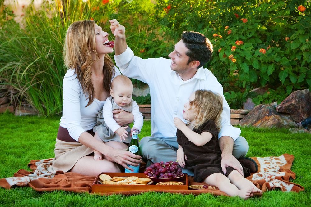 A family of four enjoys a playful moment together while sitting on a blanket in a lush green garden setting.  Photographs taken at home.