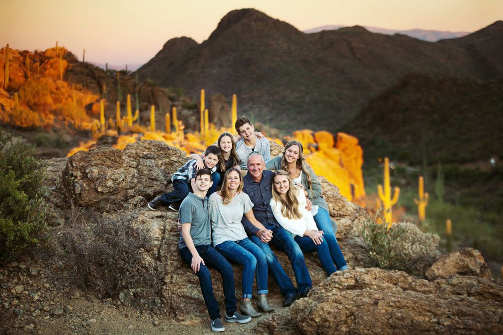 A family portrait of eight people sit on desert rocks at sunset with saguaro cacti and mountains in the background at Gates Pass in Tucson.