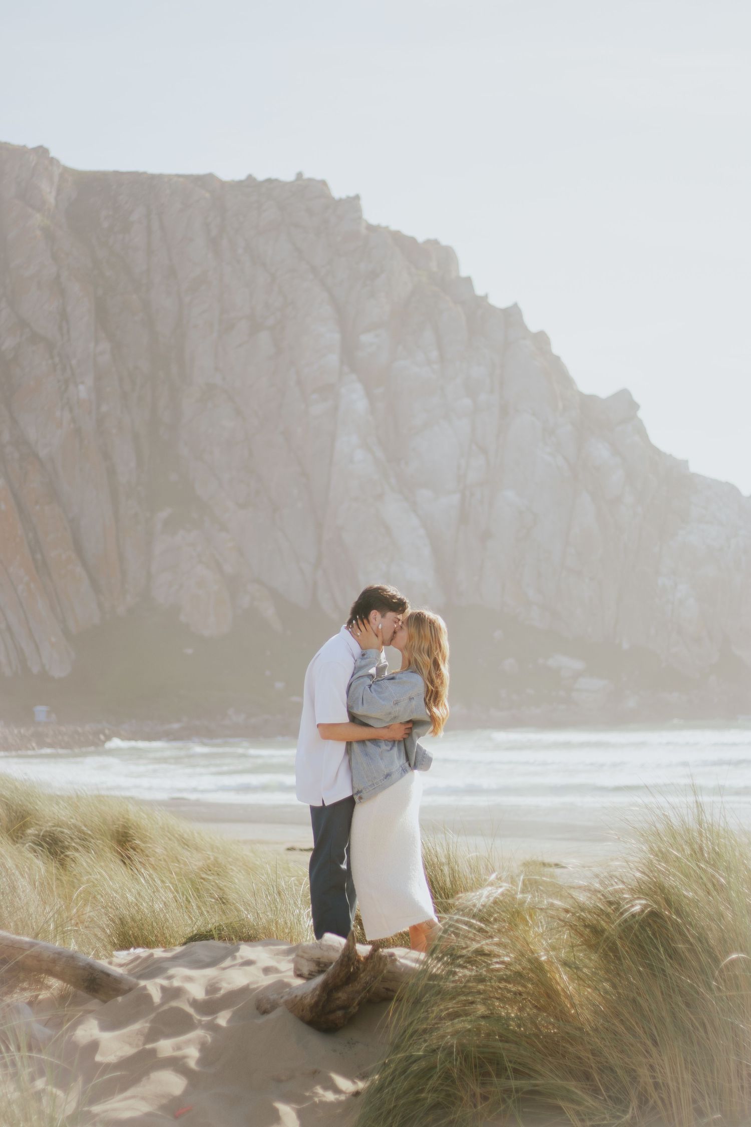 Two people share a romantic moment on a beach with tall cliffs and beach grass in the background.