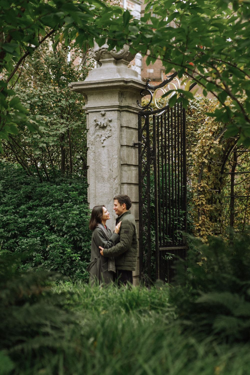 A couple stands by an ornate iron gate surrounded by greenery.