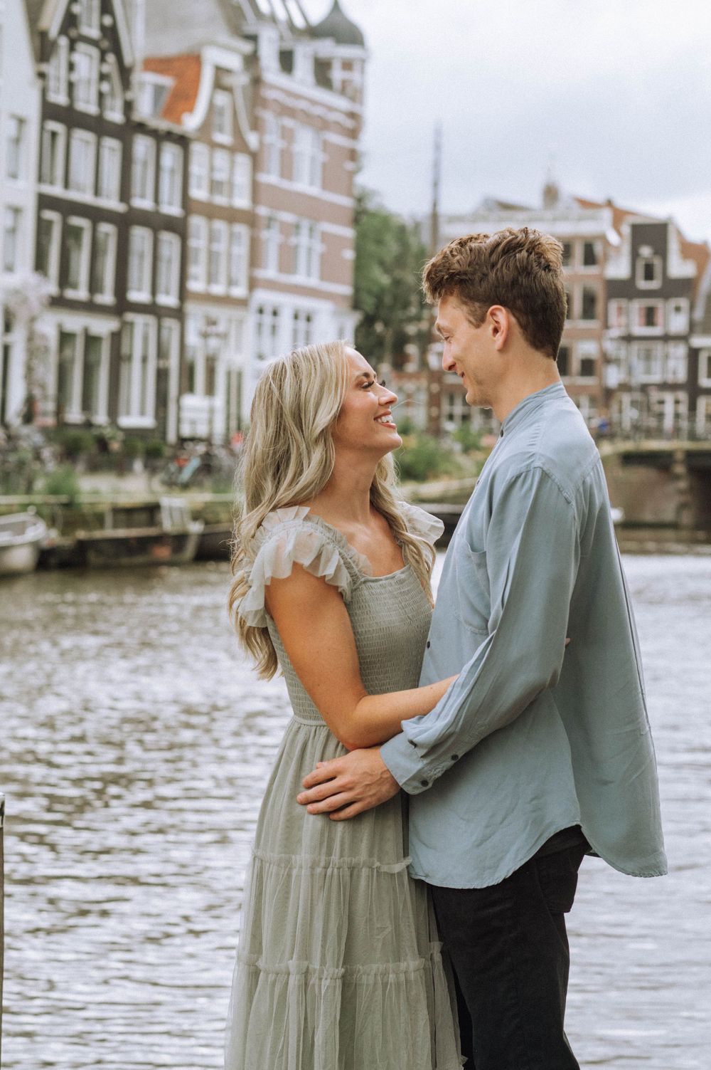 Couple embraces by Amsterdam canal with traditional Dutch architecture behind them.