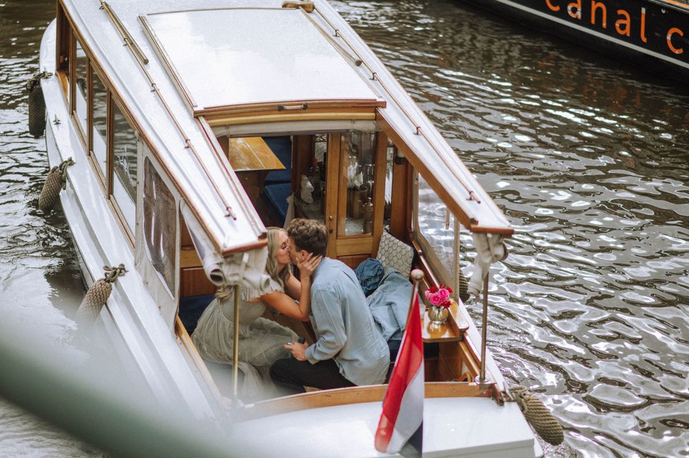 Wooden boat cruises through Amsterdam canal with historic buildings in background.