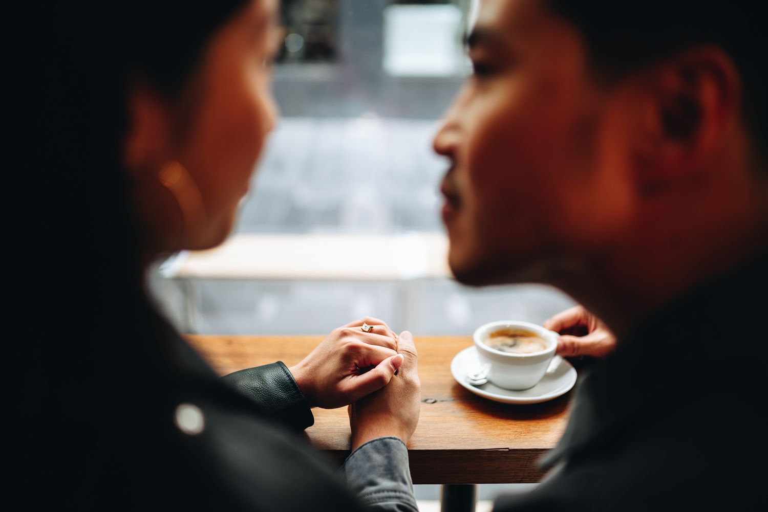 Couple holds hands across a cafe table with coffee cup in warm lighting taken by Melbourne casual couples and engagement photographer