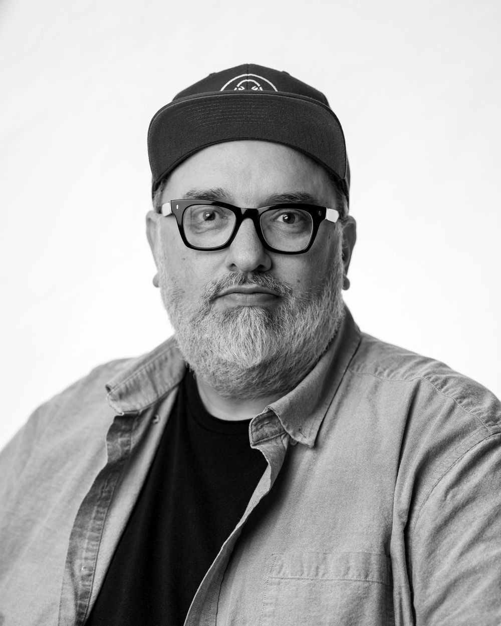 Black and white headshot of someone wearing black-rimmed glasses, a baseball cap, and a light colored jacket over dark shirt.