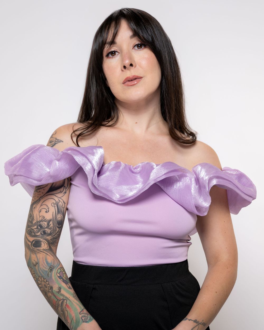 Studio portrait against white backdrop shows person in lavender off-shoulder top with ruffles and black skirt displaying arm tattoos.