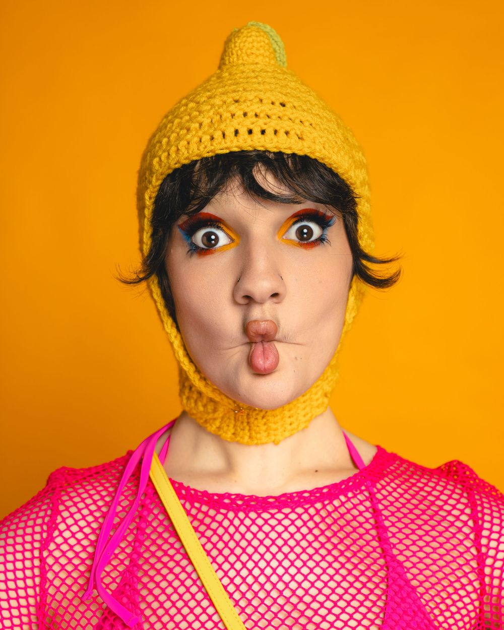 A person wearing a yellow knit hat with chin strap makes playful expression against vibrant orange background.