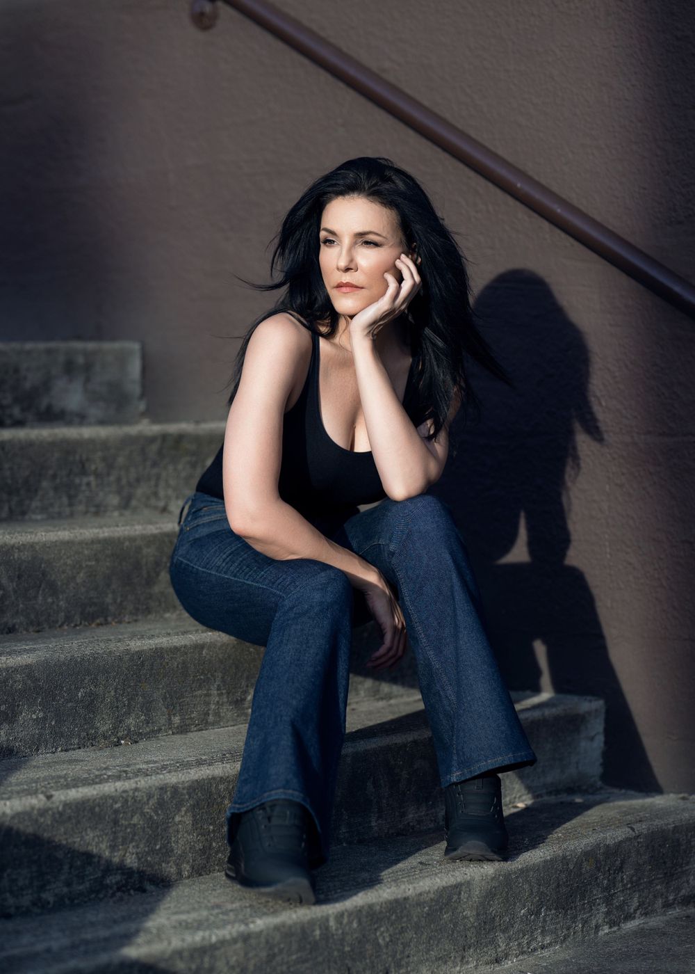 A person in dark attire sits thoughtfully on concrete steps, casting a dramatic shadow against the wall.