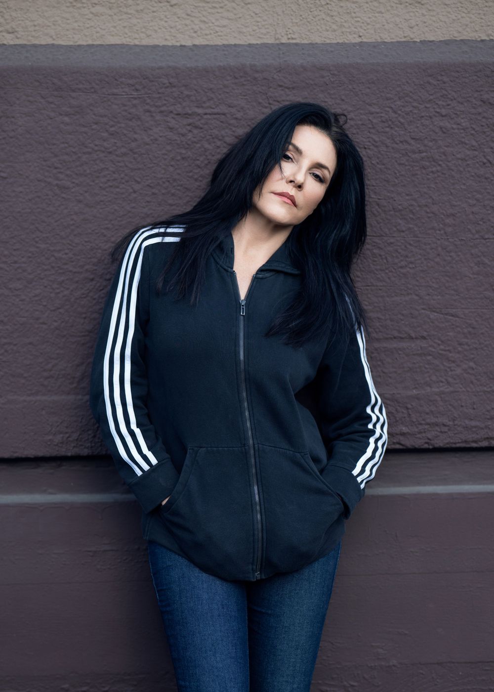 Person wearing black Adidas track jacket with white stripes and blue jeans poses against purple wall.