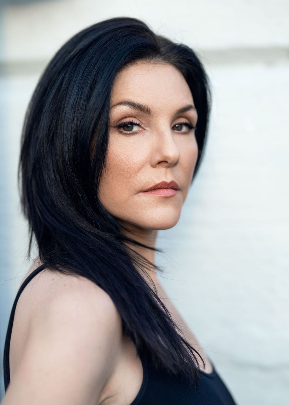 Portrait of a woman with long dark hair wearing a black sleeveless top against a light background during outdoor headshot session.