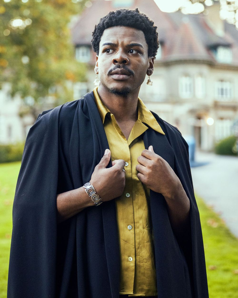 Someone poses outside wearing a yellow button-down shirt and black overcoat in front of Pittock Mansion in Portland, Oregon.