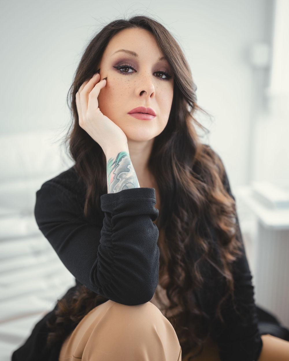 Portrait photo showing person with long wavy dark hair in black sweater and tan pants in minimalistic casual setting.