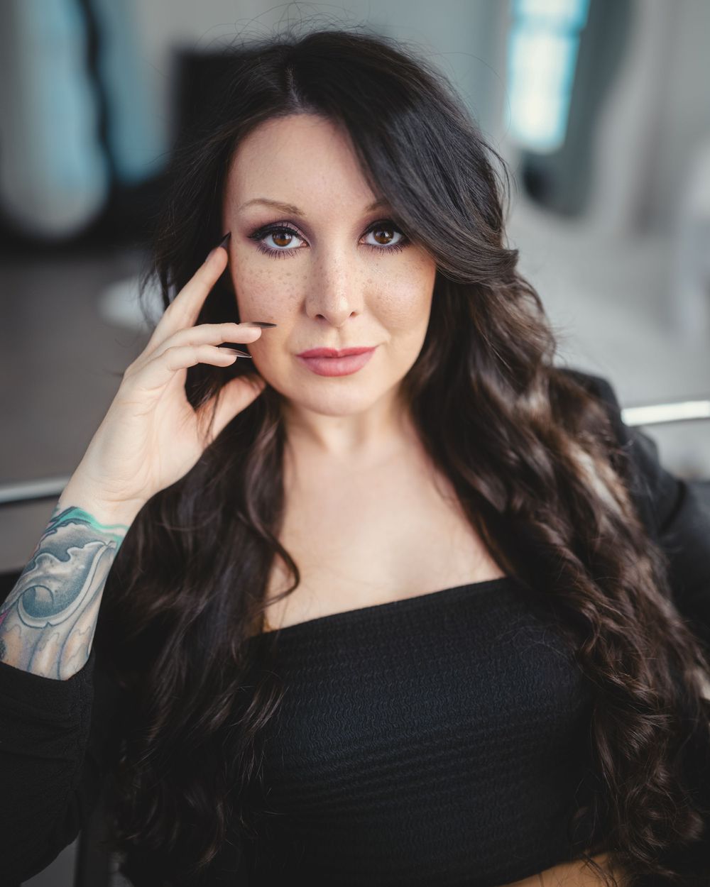 Portrait shot of a woman with long dark wavy hair wearing black dress, showing arm tattoo while touching her face.