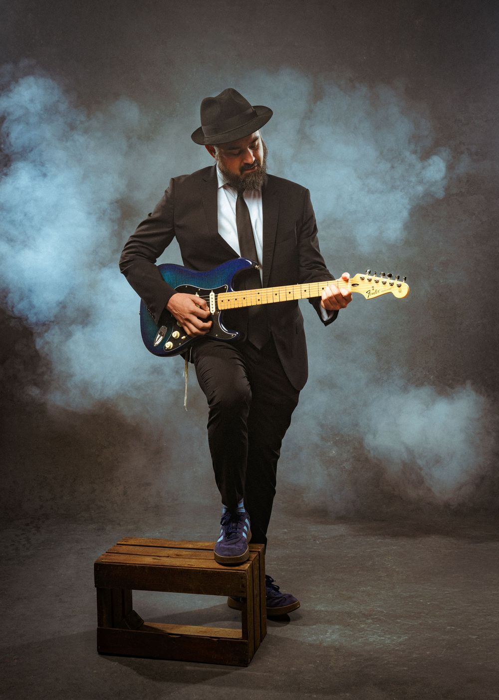 Musician in black suit and fedora playing blue Fender Stratocaster guitar on wooden box with dramatic smoke background.