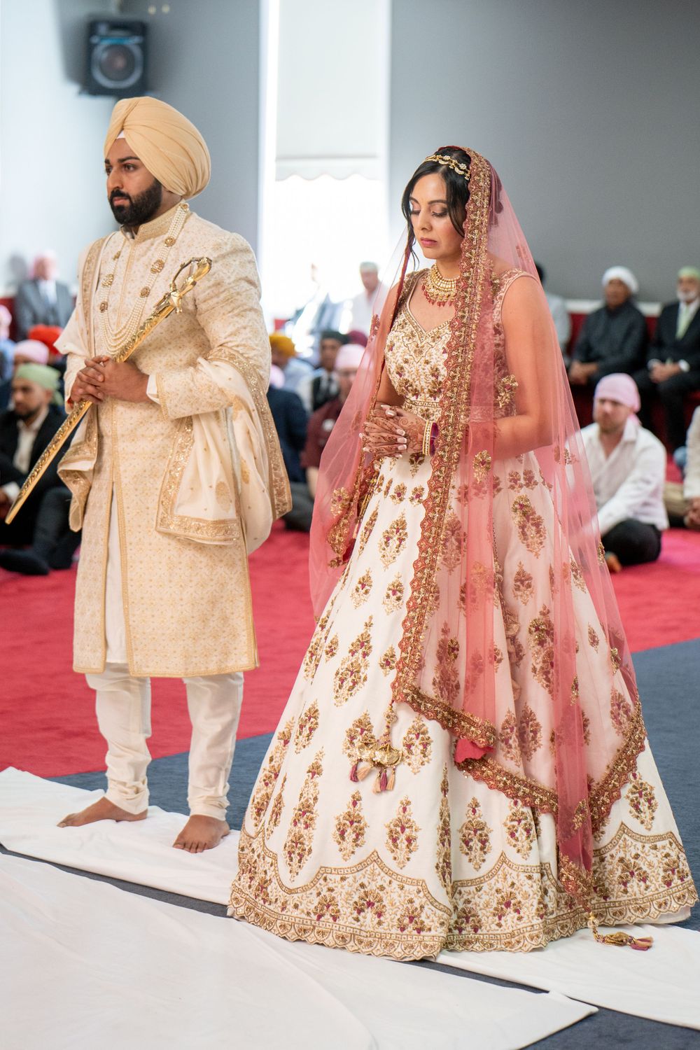 Traditional Sikh wedding ceremony with couple in ornate cream and pink wedding attire on red carpet.