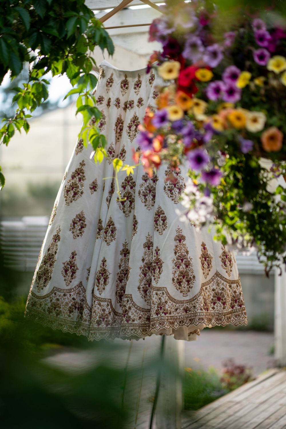 An elegant white and gold embroidered wedding lehenga dress hangs beautifully against a backdrop of colorful flowers.