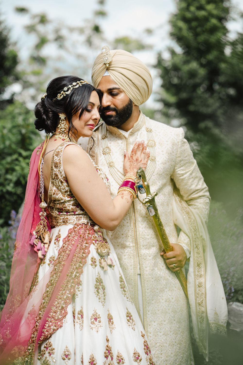 A romantic wedding portrait captures a couple in traditional Sikh and Indian attire against a green garden backdrop.