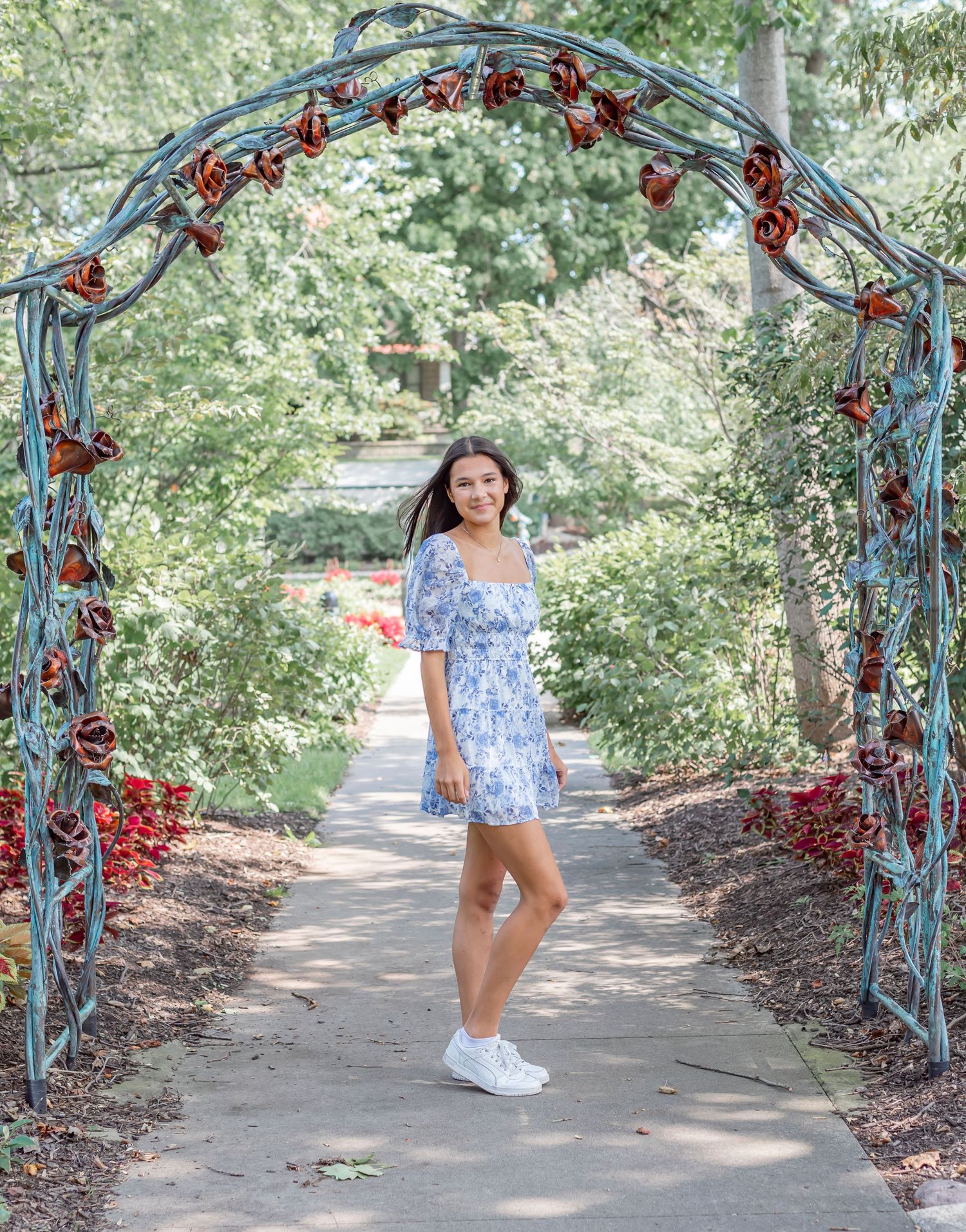 A decorative garden arch with red flowers frames a pathway through green foliage.