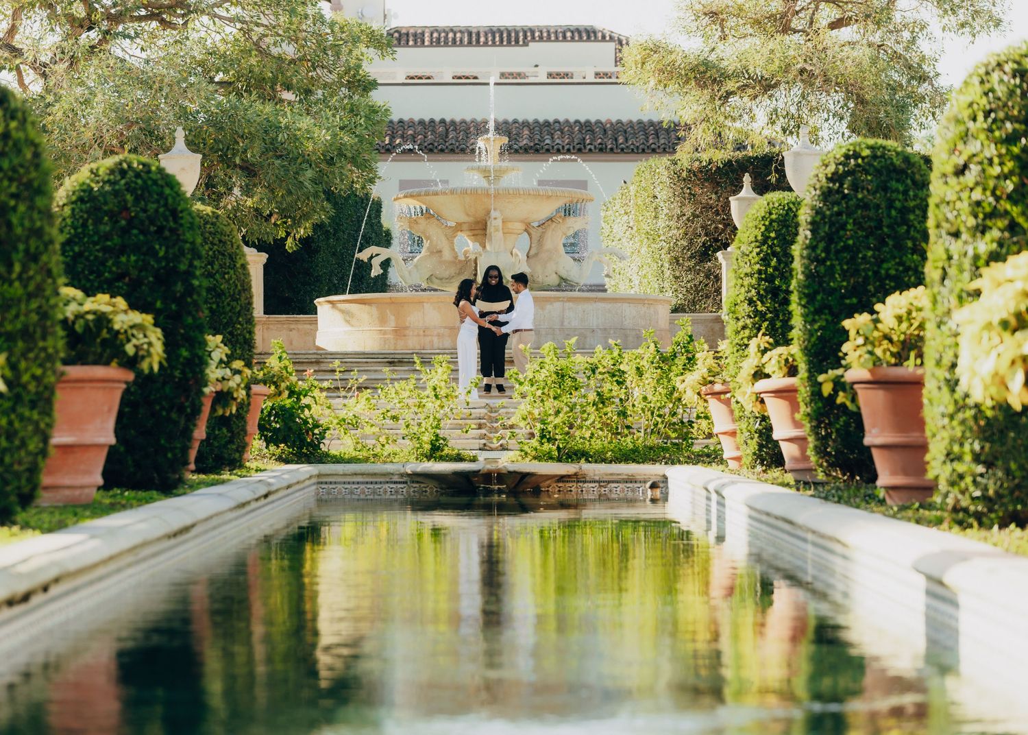 A reflecting pool leads to a classical fountain in a manicured garden with trimmed hedges.