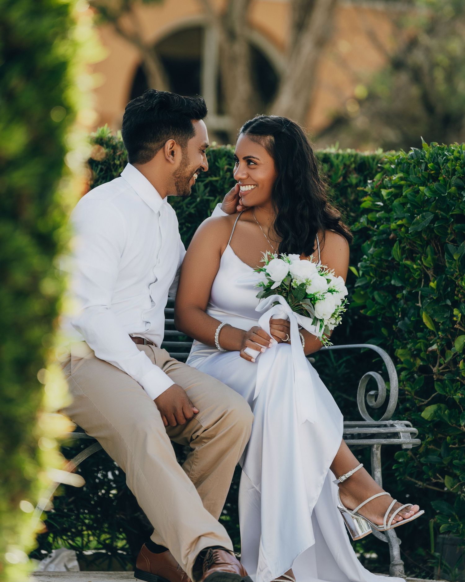 Casual wedding couple shares joyful moment on garden bench with white flowers.