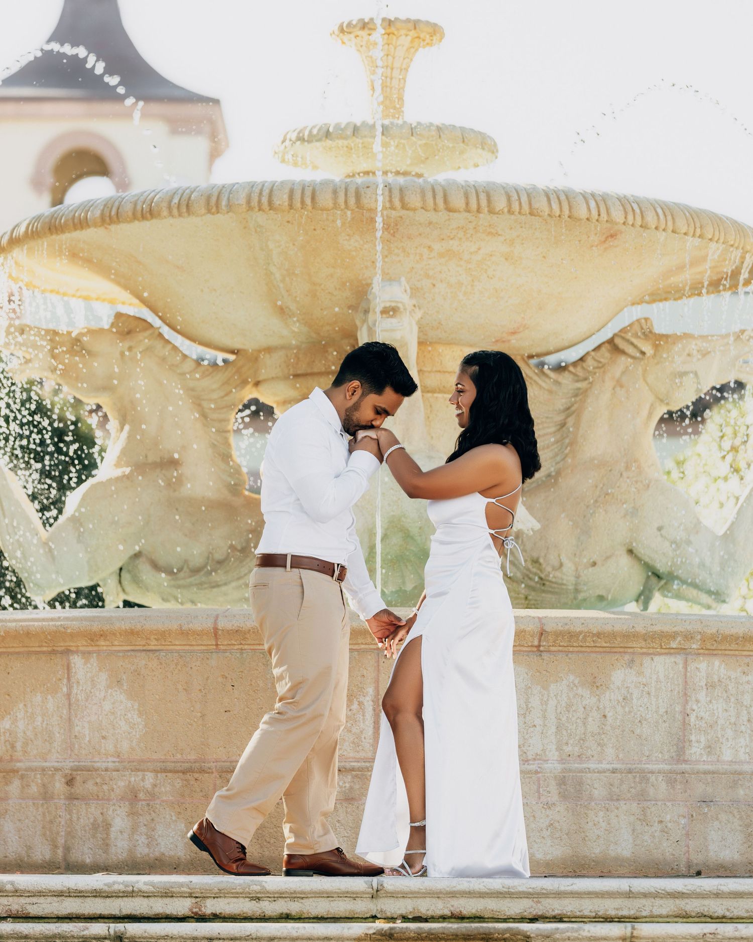 A couple shares an intimate moment by an ornate stone fountain in a romantic outdoor setting.