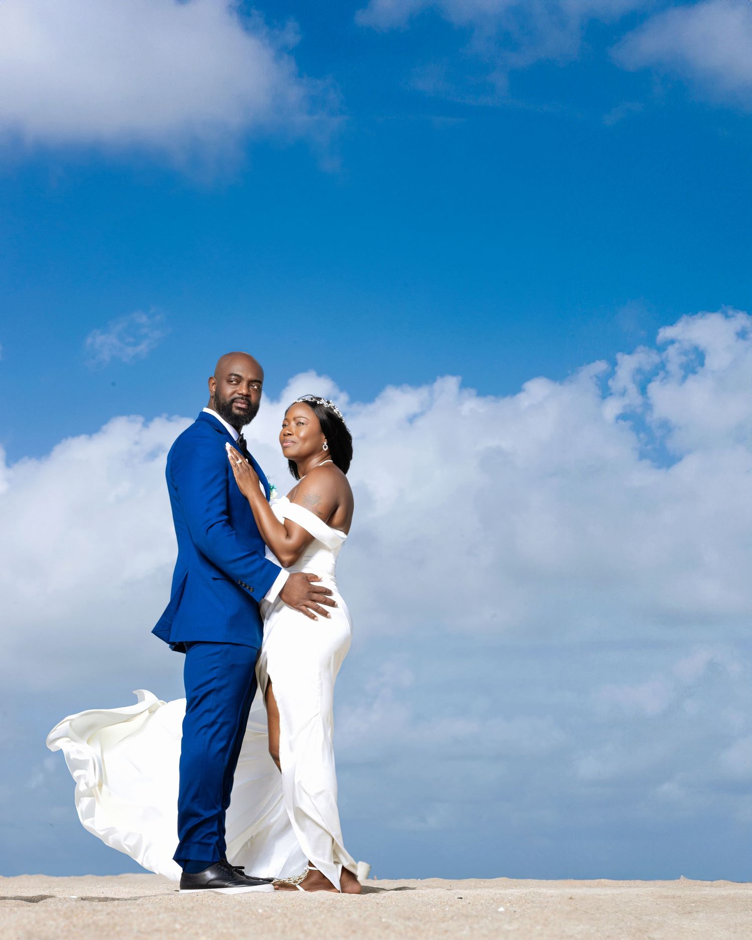 A couple in wedding attire shares an intimate moment against a bright blue sky with white clouds in the background.