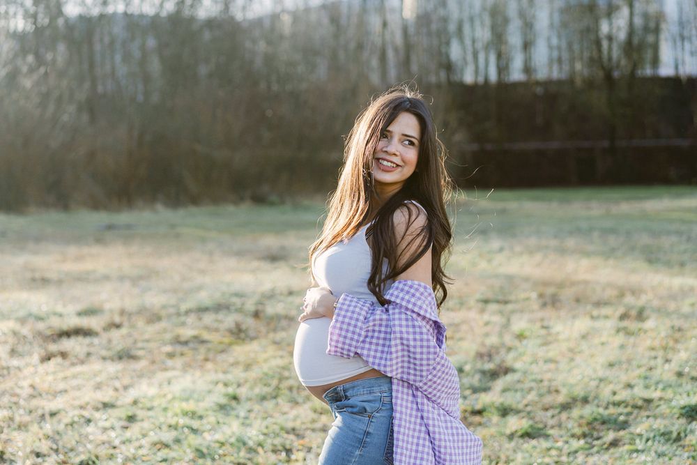 A maternity photoshoot in a field at sunset featuring a woman in jeans and white top with a purple plaid shirt.