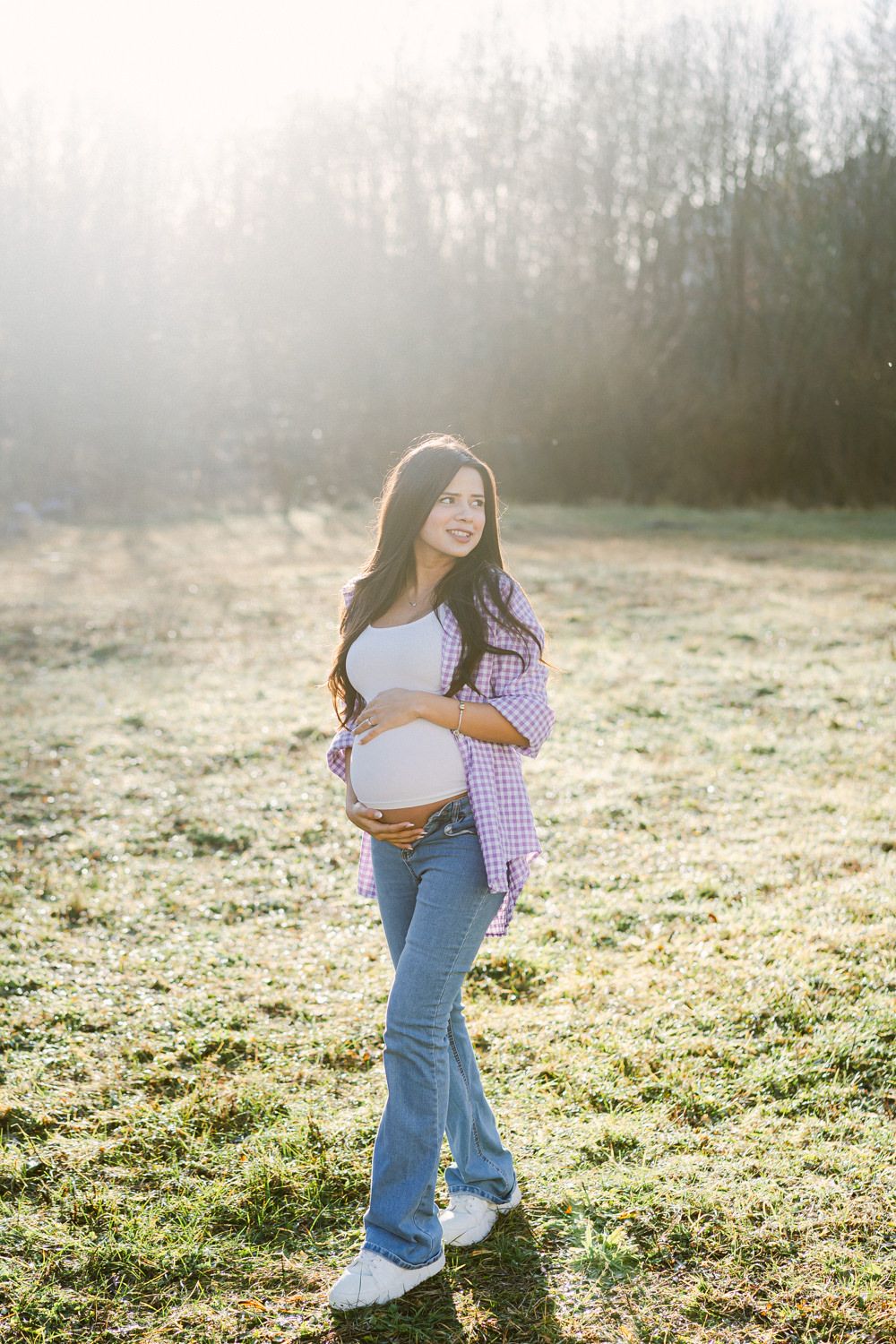 A maternity photo in a casual purple gingham shirt and jeans taken in a misty field at sunrise.